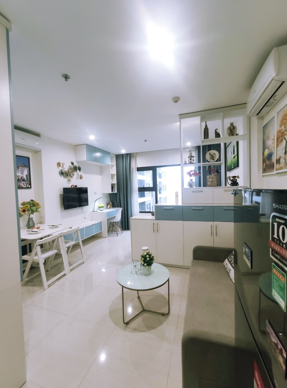 A modern studio apartment features a bright living area with sleek cabinetry and a comfortable sofa. A small glass table is adorned with a decorative flower arrangement. The dining space is equipped with a light wooden table surrounded by white chairs, complemented by ample natural light from large windows.