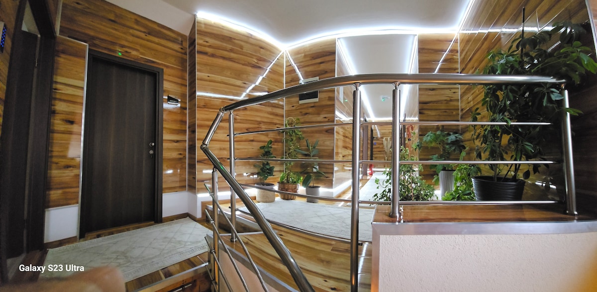 An interior hallway is featured, characterized by wooden walls and decorative greenery. Soft LED lighting creates a serene ambiance along the ceiling, while a metal railing outlines the staircase leading to an upper level.