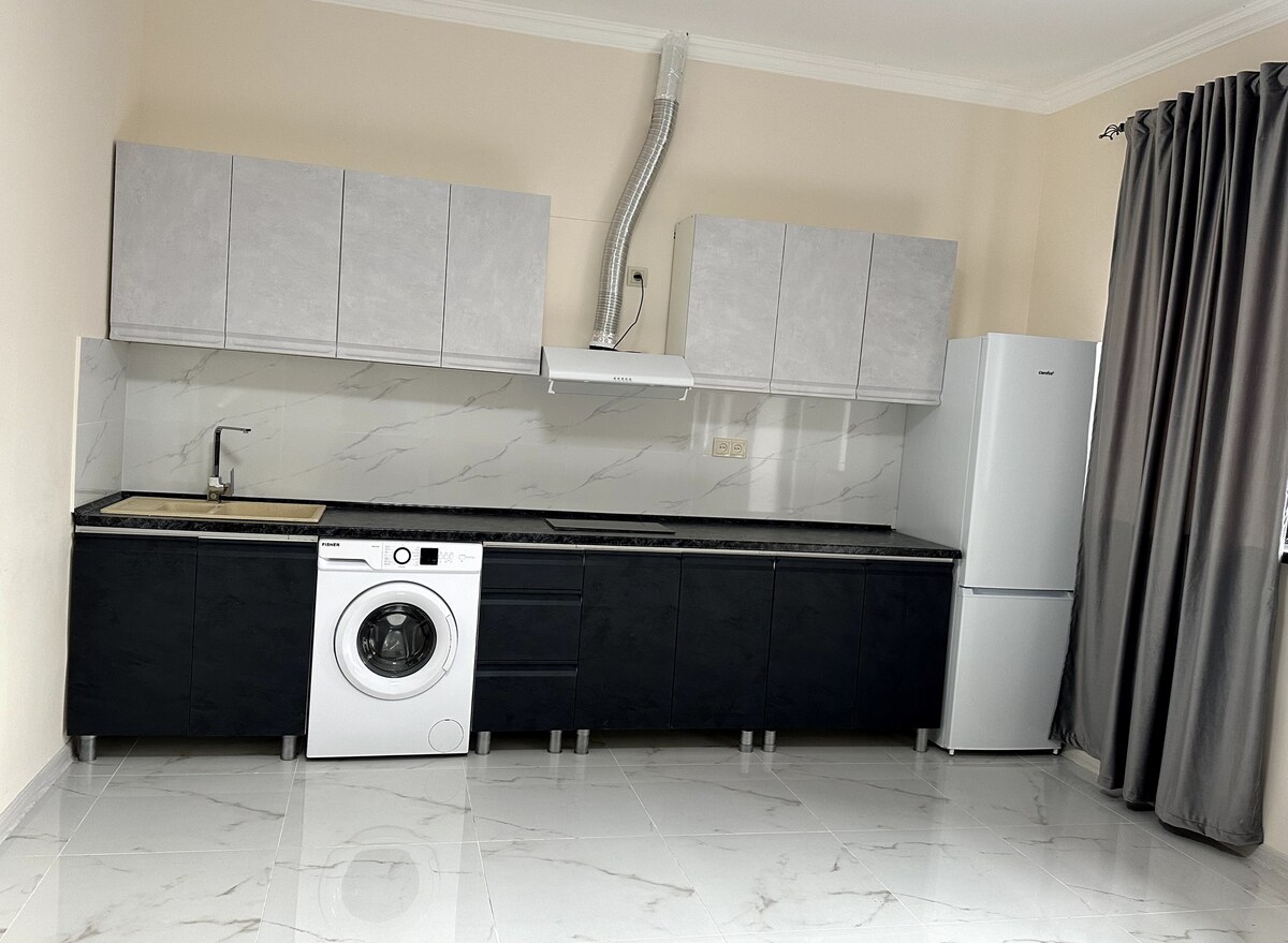 A modern kitchen features dark cabinets and a white countertop. An integrated washing machine is situated beneath the counter, with a stainless steel sink positioned beside it. A refrigerator stands at the end, and large windows are adorned with grey curtains, allowing natural light to fill the space.