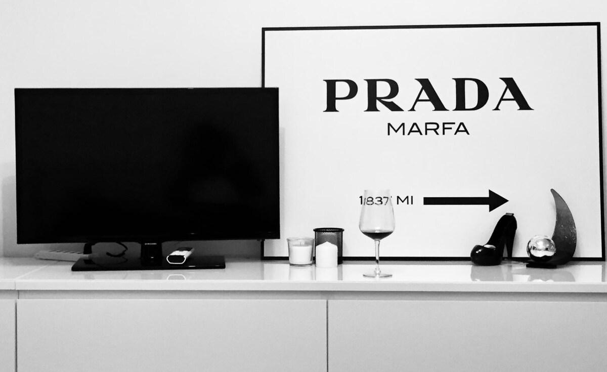 A modern TV sits atop a sleek white surface, accompanied by decorative items including a candle, a wine glass, a high-heeled shoe, and a shiny orb. A large artwork with the word 'PRADA' is prominently displayed in the background.