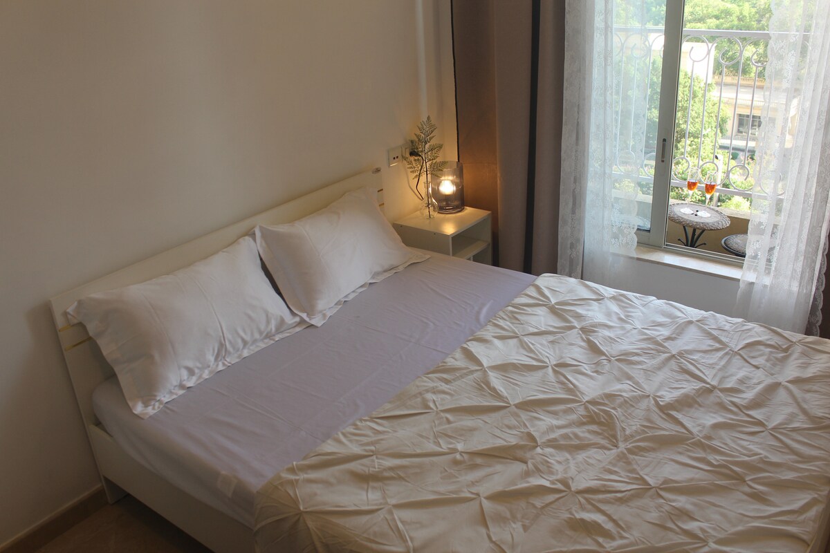 A cozy bedroom showcases a plush bed with white linens and a quilted cover. Natural light filters through sheer curtains, illuminating the space. A bedside table holds a softly glowing lamp, complementing the tranquil atmosphere. A window offers a view of lush greenery outside.
