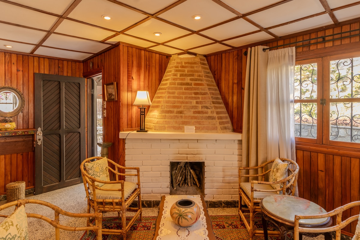 The living area features a rustic design with wooden walls and a central brick fireplace. Two wicker chairs are positioned around a small table, while warm lighting from a lamp enhances the cozy ambiance. A window with decorative ironwork allows natural light to fill the space.