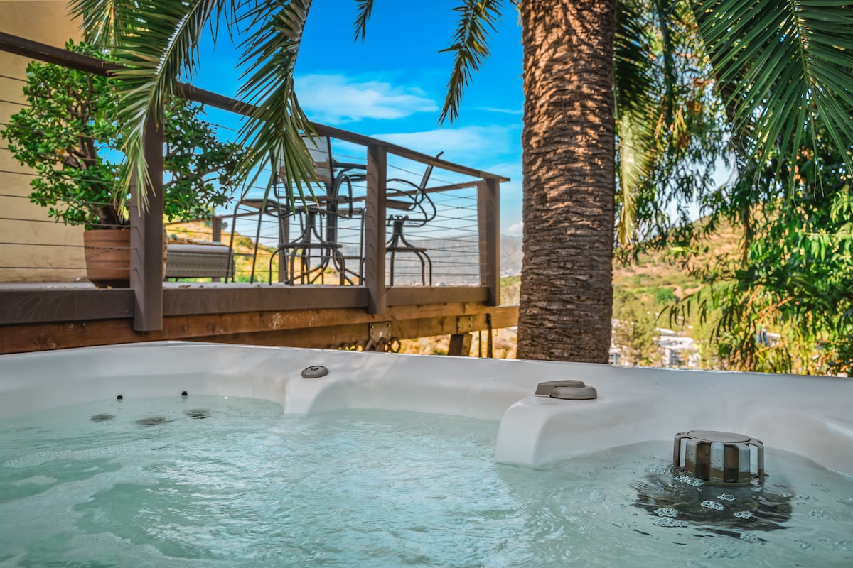 A hot tub is set on a wooden deck, surrounded by palm trees and greenery. In the background, a dining set with chairs is visible, offering a view of the hills. Clear skies and distant landscape are seen, enhancing the outdoor setting.
