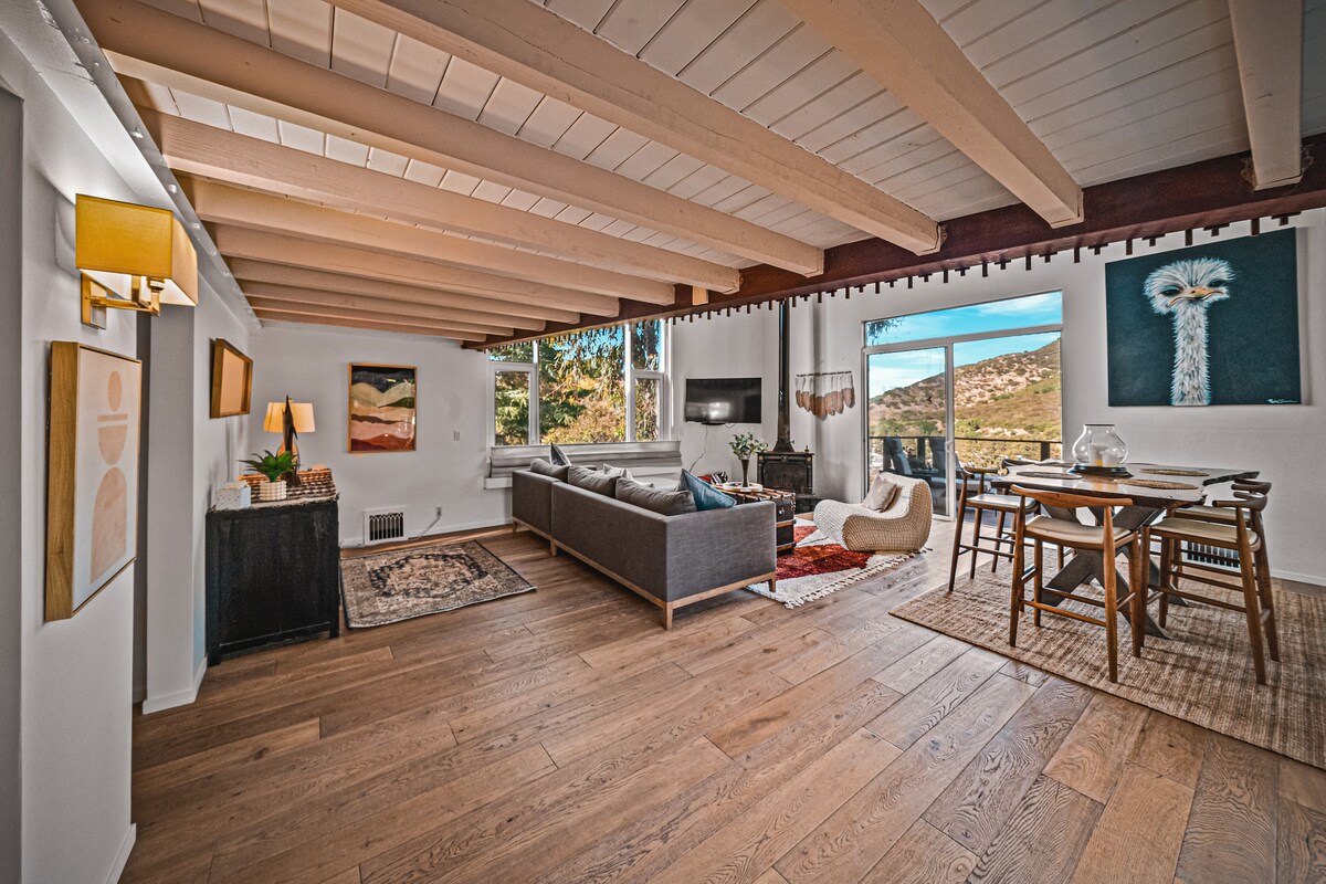 An open living area is illuminated by natural light, featuring a comfortable seating arrangement with a sectional sofa. A dining table is positioned nearby, with artwork adorning the walls. Large sliding doors lead to an outdoor space, enhancing the connection between indoor and outdoor living.