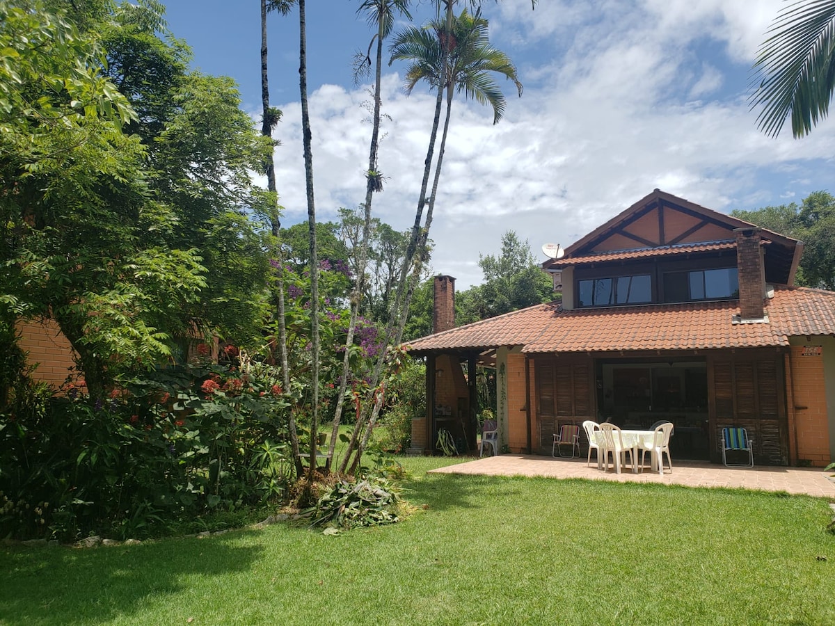 A two-story house is set within a lush garden, surrounded by tall trees and vibrant foliage. The outdoor space includes a grassy area, and a covered patio with light-colored furniture. The home's wooden exterior and tiled roof contribute to its cozy appeal.