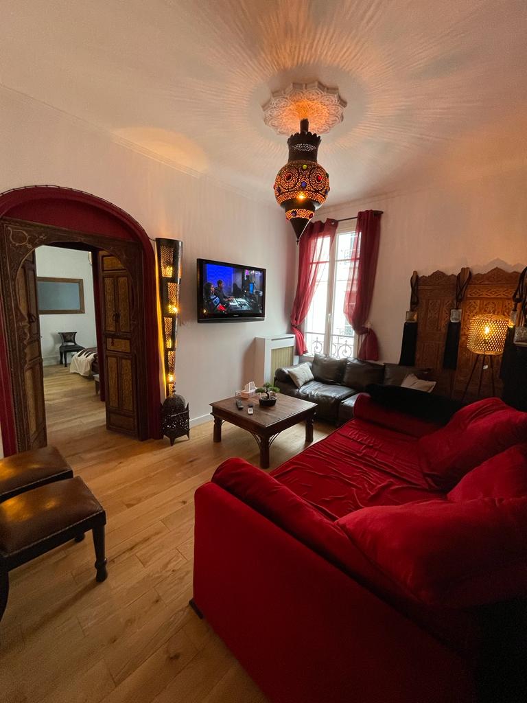 A living area is highlighted by a red sofa and wooden chairs, with decorative lanterns contributing to the ambiance. A television is mounted on the wall, providing entertainment options. Natural light enters through a window adorned with sheer curtains, enhancing the cozy atmosphere.