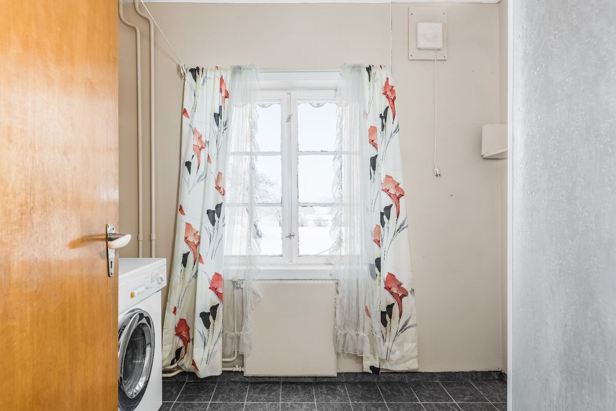 A laundry area is showcased, featuring a washing machine alongside a window adorned with floral-patterned curtains. Natural light enters through the window, illuminating the neutral wall colors and dark tiled floor.