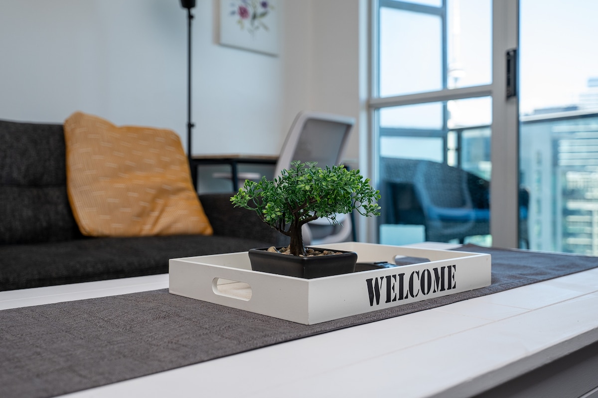 A welcoming living area is highlighted by a low coffee table with a decorative tray inscribed with 'WELCOME.' A small potted plant adds a touch of greenery. The background features a comfortable couch, a modern chair, and floor-to-ceiling windows with city views.