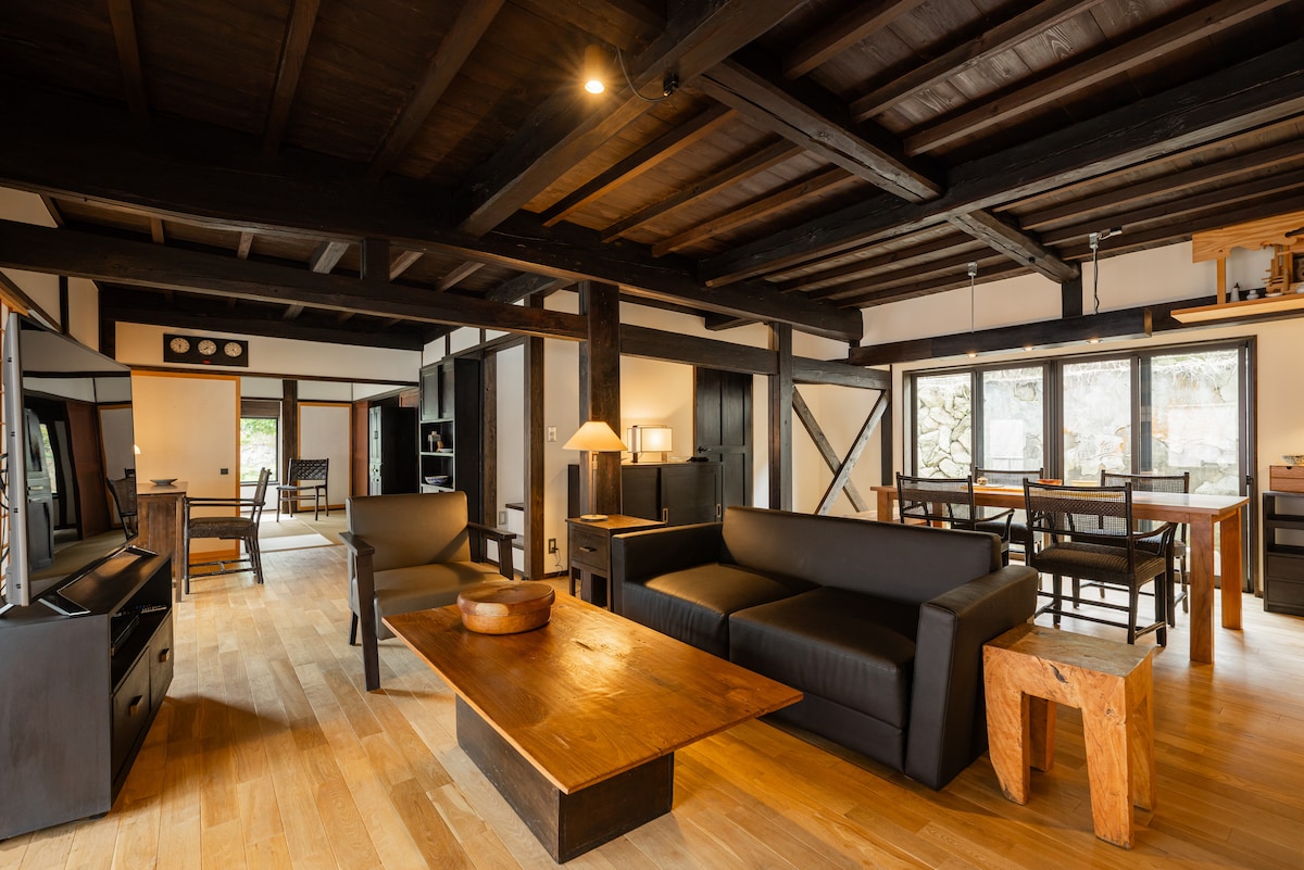A spacious living area features dark wood beams and modern furnishings. A comfortable sofa faces a large coffee table, complemented by a light fixture. Large windows provide natural light and views of the surrounding nature, while a dining table and chairs are positioned nearby.