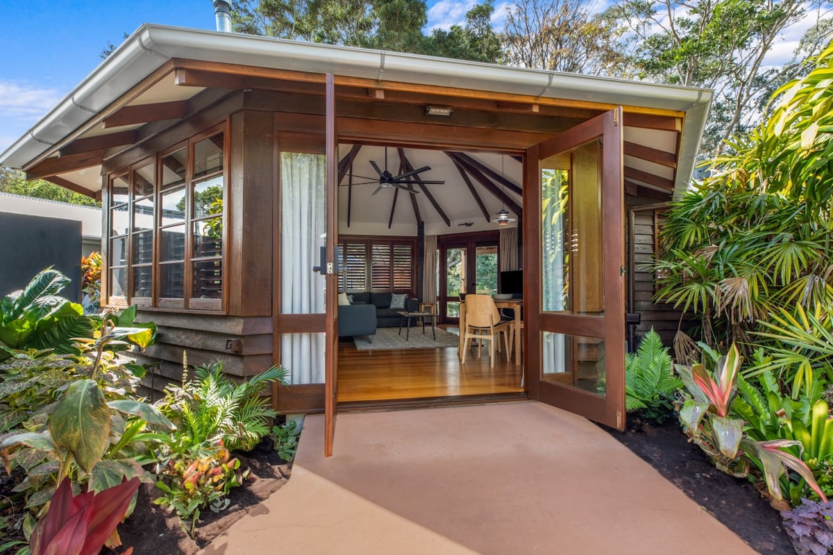 The entrance to a hexagonal villa is framed by lush greenery and vibrant plants. Large wooden doors open to reveal an indoor living space with wooden flooring and high ceilings. Natural light filters through large windows, connecting the indoor and outdoor environments.
