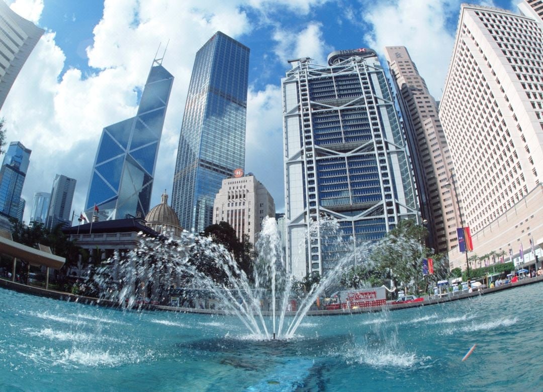 A vibrant urban scene featuring a modern fountain at the forefront, surrounded by towering skyscrapers and blue skies. The dynamic architecture of glass and steel structures reflects the city's energy, creating a striking backdrop for the lively water display.