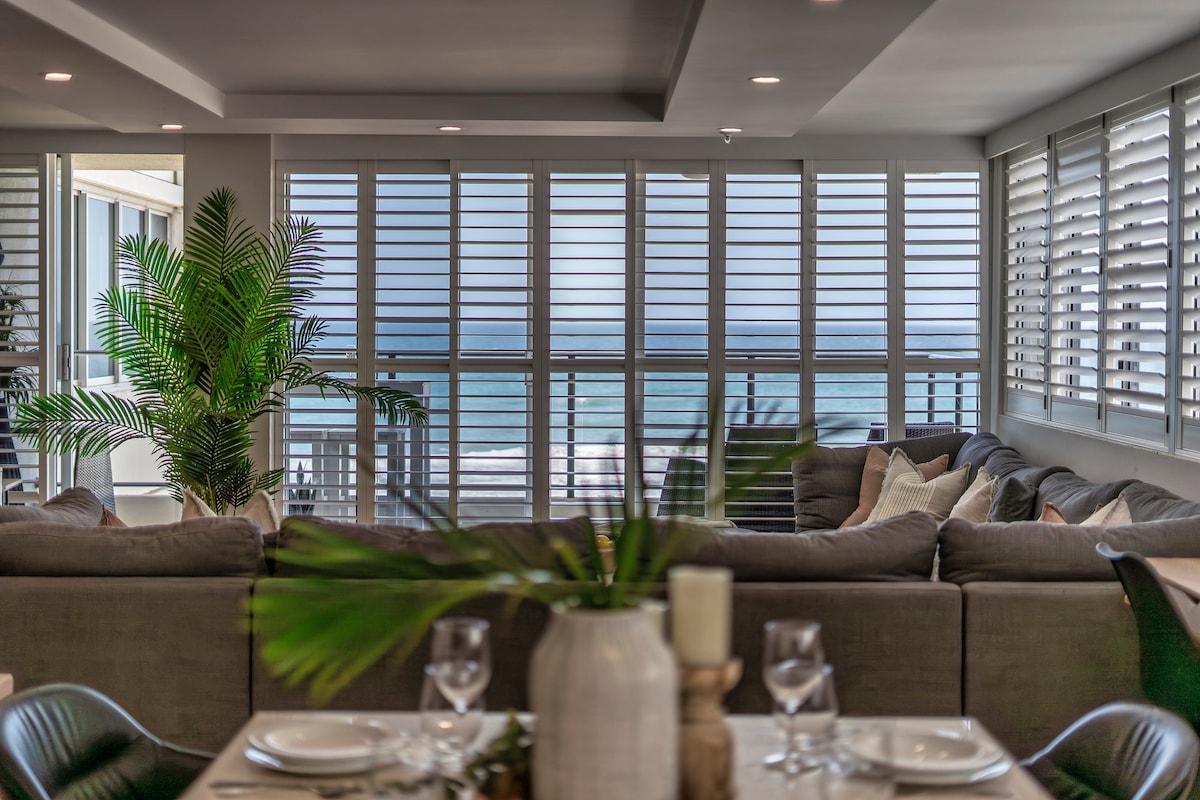 A spacious living area is showcased with large windows adorned with shutters, allowing glimpses of the ocean. Comfortable seating is arranged around a central coffee table, complemented by indoor plants and a dining setup in the foreground, inviting relaxation and social gatherings.