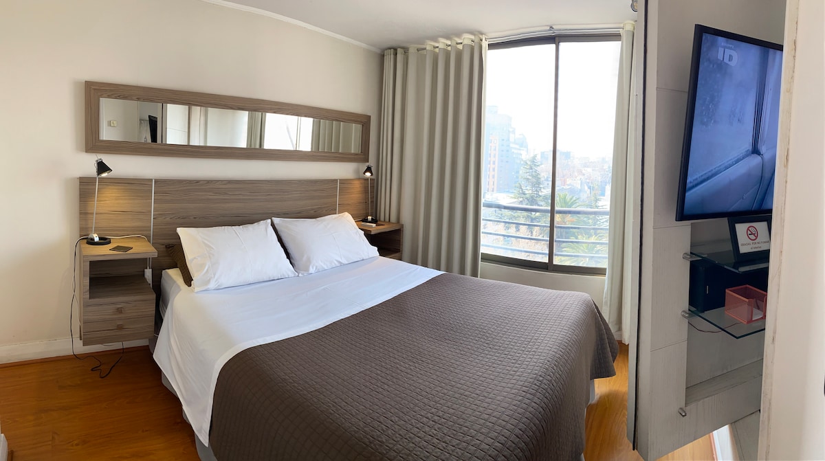 A well-lit bedroom features a spacious bed with soft linens and a neutral-colored blanket. A large window provides views of the surrounding area, while a TV is mounted on the wall opposite the bed. Two bedside tables with lamps are positioned on either side.
