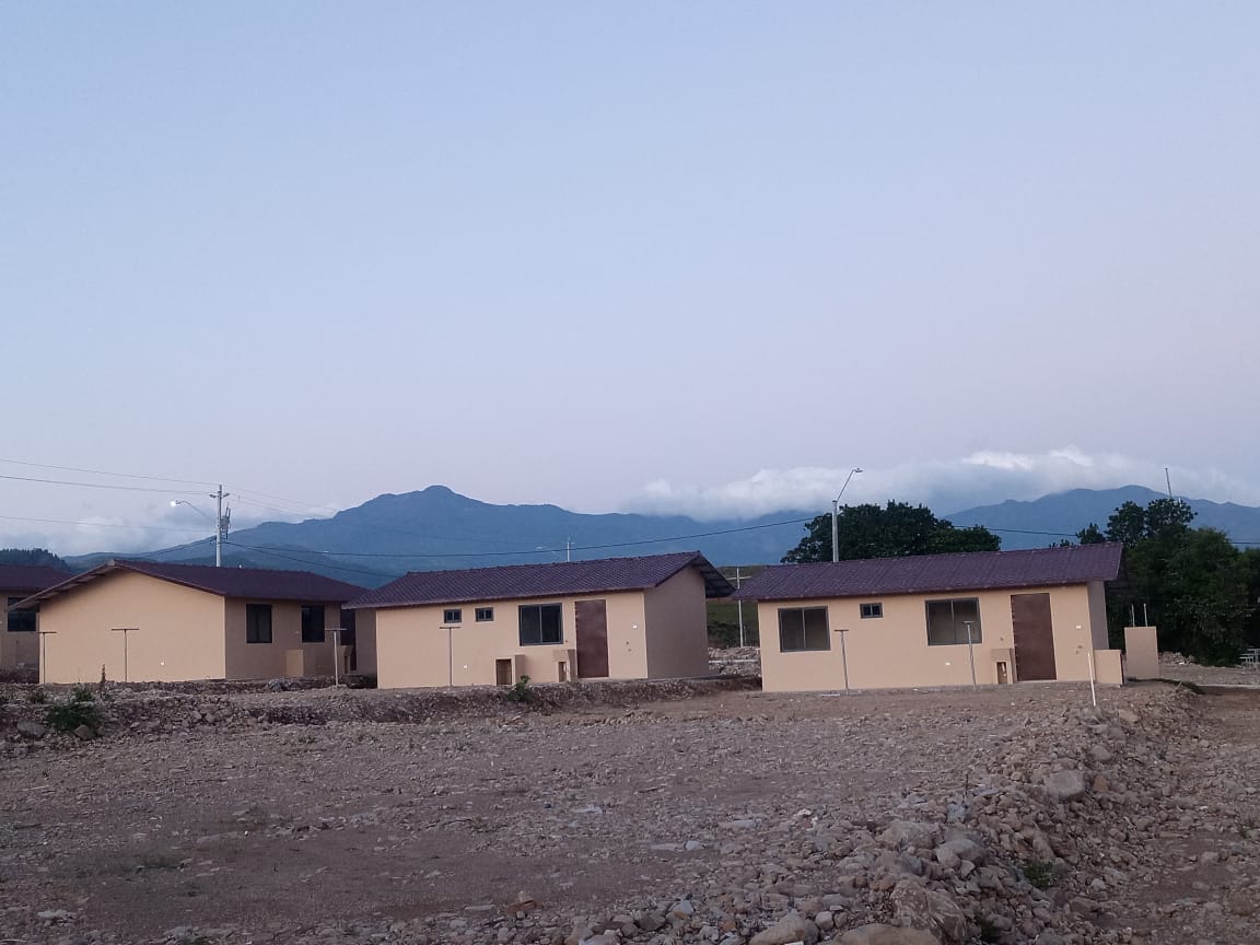 A row of single-story buildings is situated against a mountain backdrop, with gentle hills and clouds in the distance. The structures feature muted tones and simple design, providing a sense of serenity in a natural environment.