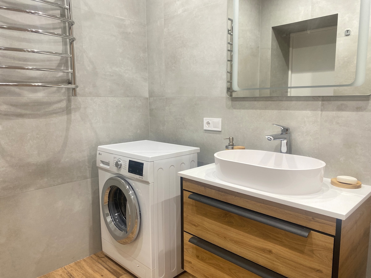 A modern bathroom is presented with gray tiled walls and a wooden vanity. A white washing machine is positioned beside the basin, which features a round vessel sink. A large mirror hangs above the sink, and a heated towel rack is installed on the wall.