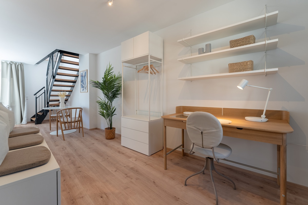 A modern living area features a wooden desk, chair, and shelving unit. A potted plant adds a touch of greenery, while a staircase leads to the upper level. Natural light fills the space, enhancing the open layout with comfortable seating and a simple decor.