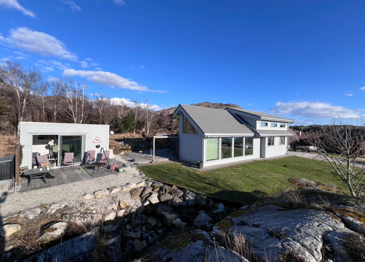 A contemporary cabin is set against a clear blue sky, featuring large windows that invite natural light. The well-maintained outdoor area includes a patio with seating and a lawn bordered by rocky terrain and trees, creating a serene and natural setting.