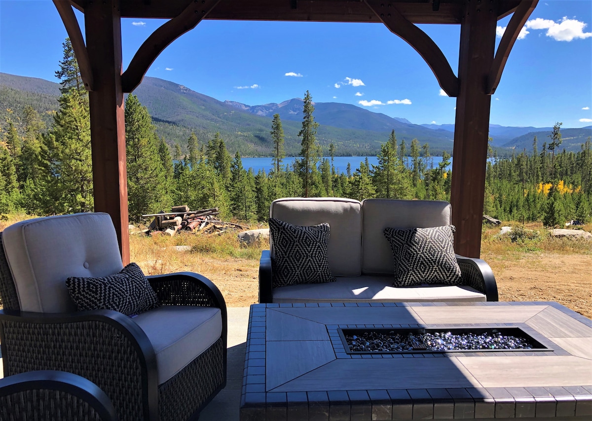 An outdoor seating area is situated under a wooden pergola, featuring two cushioned chairs and a sofa with neutral tones and decorative pillows. A gas fireplace table is positioned at the center, offering views of the expansive landscape, including mountains and a lake in the background.