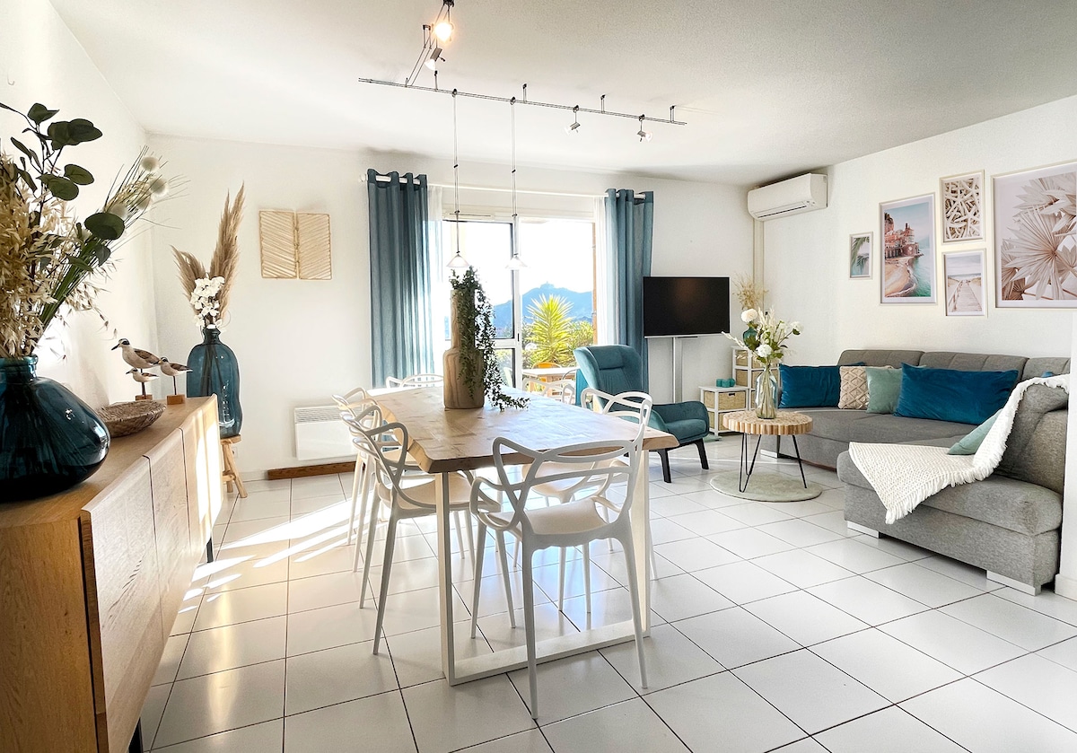 A bright living space features a large dining table surrounded by white chairs. A comfortable sofa is positioned to the side, complemented by decorative cushions. Natural light floods the room through a window with sheer curtains, offering a view of nearby mountains.
