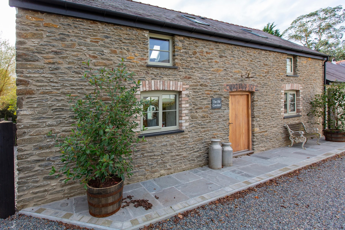 The exterior of a renovated stone cottage is displayed, showcasing a rustic facade with a wooden door framed by greenery. Paved stones lead to the entrance, complemented by two large planters and a decorative bench.