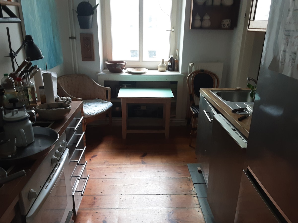 A kitchen area features a mix of light and dark wooden elements. A well-equipped stove is positioned adjacent to a sleek stainless steel refrigerator. A small table is centered in front of a window, with a cozy chair placed nearby. Soft natural light enters through the window.
