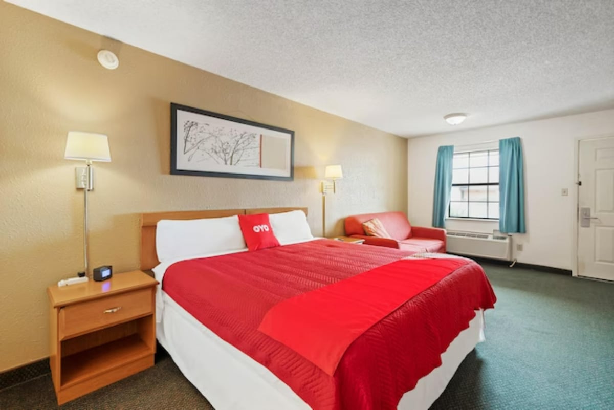 A spacious room features a large bed adorned with a red coverlet. A bedside table holds a small clock. Adjacent, a seating area with a red couch is visible. Natural light enters through the window, framed with light blue curtains.