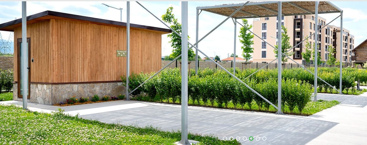 A sunshade structure featuring a wooden cabin is situated within a landscaped area. Neat hedges and greenery surround the paved path, creating a sense of openness. The sunshade is supported by steel beams, offering a shaded space and a clear view of nearby plants and the sky.