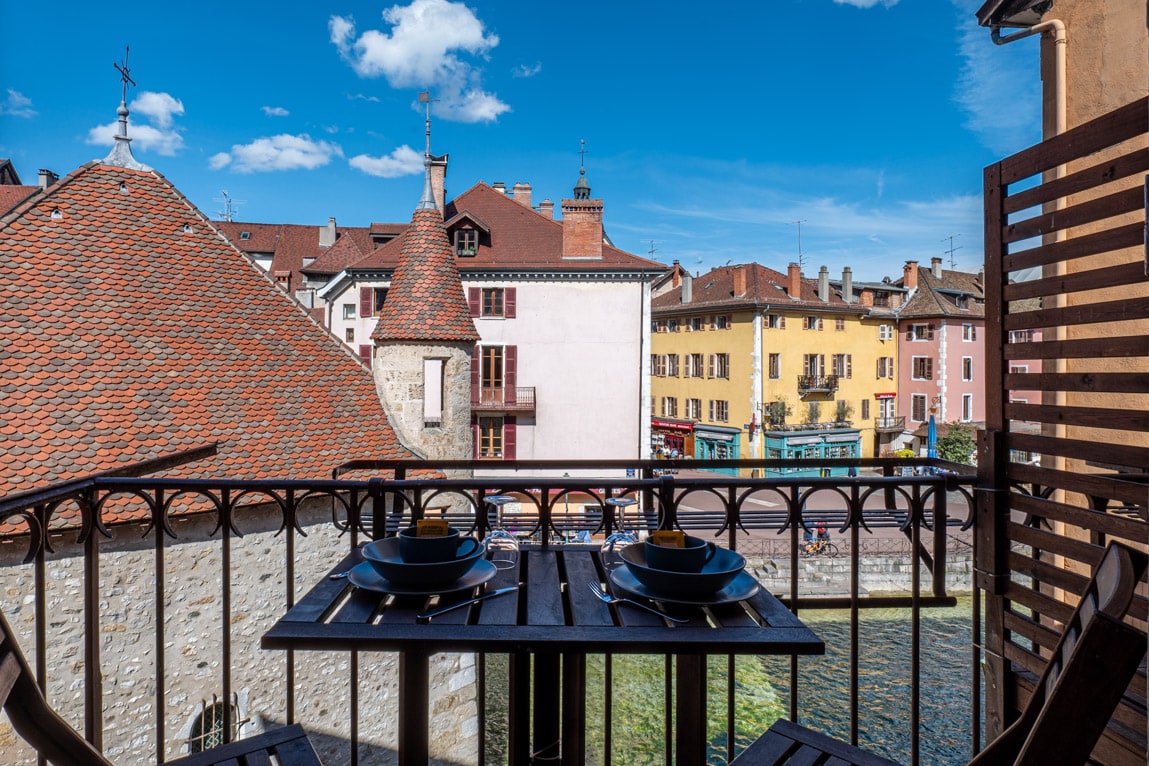 A small balcony is visible, featuring a wooden table set with two plates and utensils. The surroundings are characterized by historical buildings with varying colors, including shades of beige and yellow. A clear blue sky adds a bright backdrop to the scene.