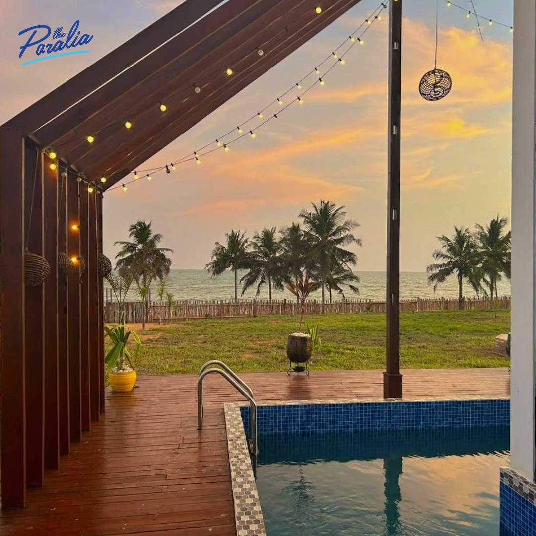 A serene outdoor space features a private pool framed by wooden decking. Soft lighting hangs overhead, casting a warm glow. The view includes palm trees swaying gently against a colorful sunset sky, leading towards the tranquil sea in the background.