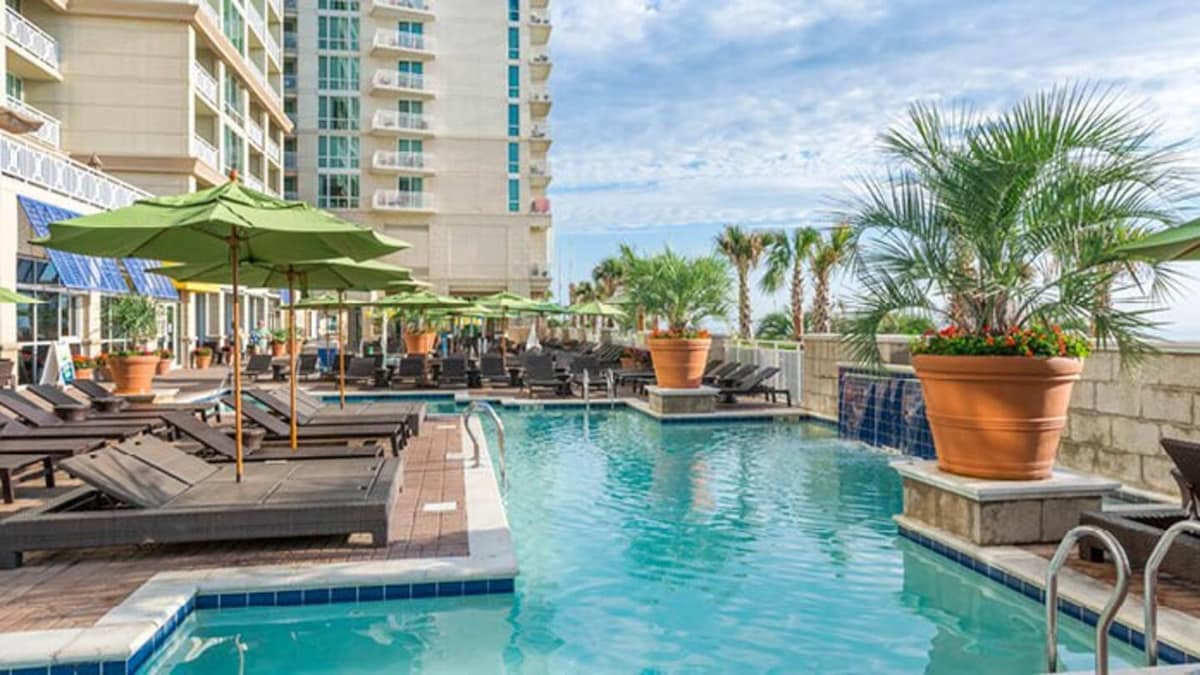 A refreshing outdoor pool is framed by lounge chairs under green umbrellas. Potted palm trees and colorful flowers add a vibrant touch around the pool area. The building in the background features large windows, suggesting upscale accommodations and a lively atmosphere.