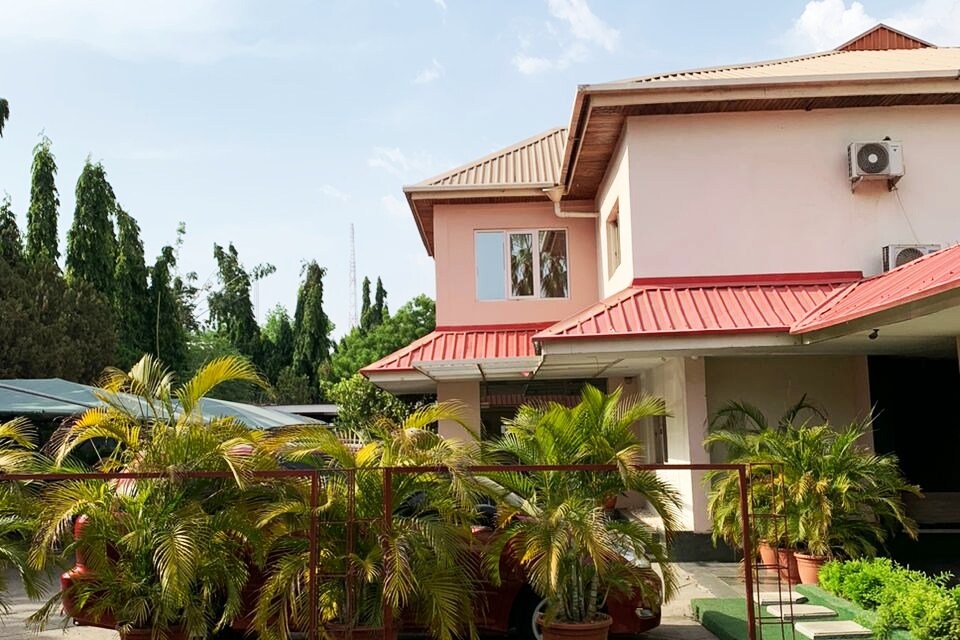 A two-story building with a red roof is framed by lush greenery and potted plants at the entrance. Large windows reflect natural light, creating an inviting facade. The exterior is complemented by a well-maintained pathway leading to the main door.
