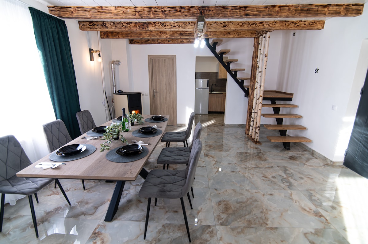 A dining area includes a large wooden table set for six, accompanied by upholstered chairs. Natural light filters through the windows, highlighting the marble flooring. A staircase is visible in the background, leading to the upper level, while a kitchenette is subtly integrated on the side.