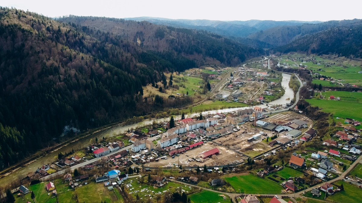 An aerial view captures a serene landscape featuring a valley surrounded by rolling hills and mountains. A small town is nestled along a river, with houses of various colors dotting the area. Green fields extend into the distance, creating a peaceful rural scene.