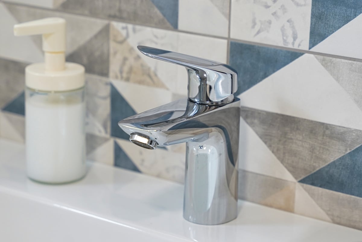 A modern faucet is positioned on a white sink, reflecting light. A transparent soap dispenser sits nearby, featuring a simple design. The backdrop displays a wall with geometric tiles in blue, cream, and grey hues, adding a stylish touch to the bathroom.
