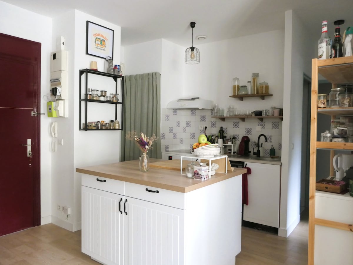 A well-equipped kitchen is showcased, featuring a central wooden island with seating and a variety of appliances. Open shelves display glass jars and cooking essentials. The space is illuminated by natural light, complemented by white cabinetry and tiled backsplash, creating a functional and inviting cooking environment.