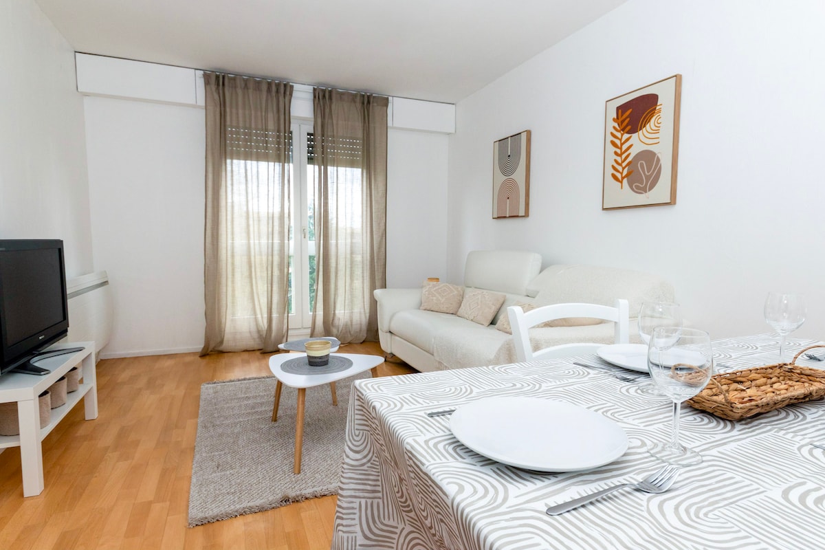 A bright living area features a comfortable white sofa paired with a stylish coffee table. Light wooden flooring and large windows with sheer curtains allow natural light to fill the space. Wall art complements the neutral decor, enhancing the calming ambiance.