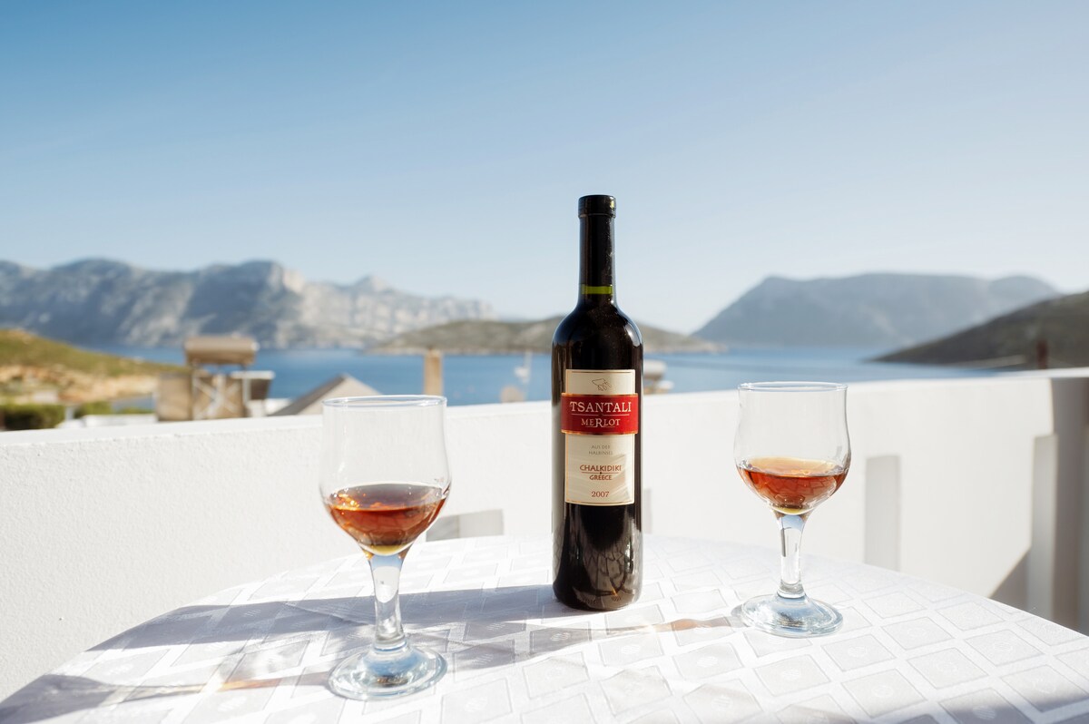 A bottle of Tsantali Merlot is showcased on a table, flanked by two glasses filled with wine. The backdrop features a serene view of the Aegean Sea and surrounding hills under clear blue skies, creating a peaceful atmosphere for relaxation.