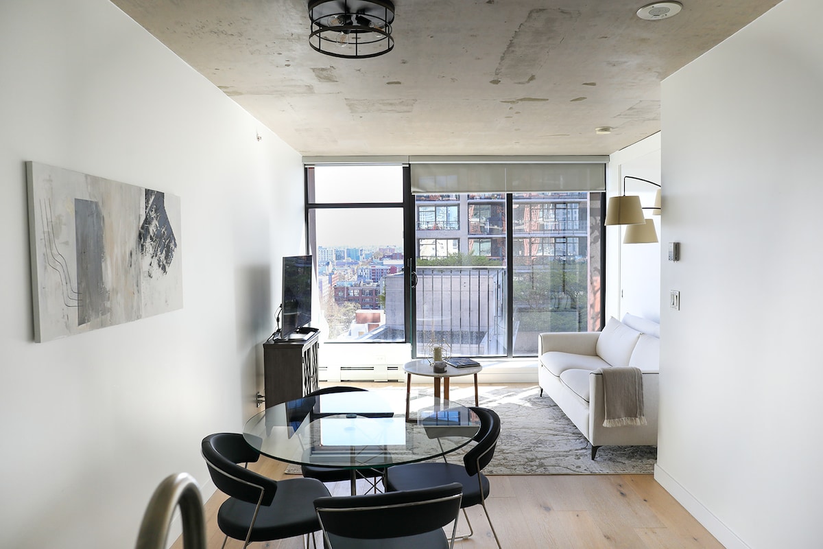 The living area features floor-to-ceiling windows providing abundant natural light and views of the city. A modern glass dining table is surrounded by four sleek black chairs. A comfortable sofa is placed against the wall, complemented by minimalist decor.