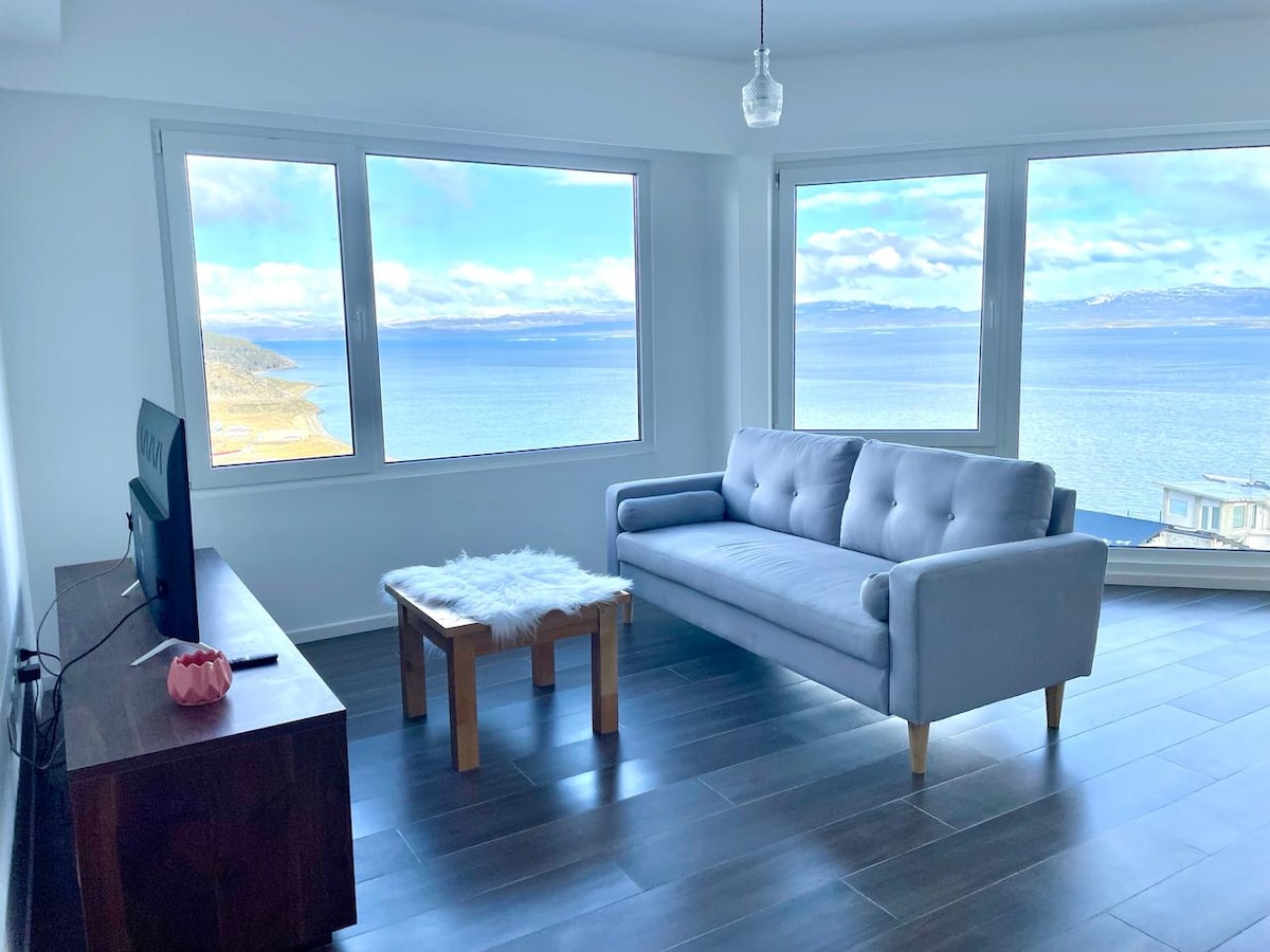 A bright living area is highlighted by large windows framing scenic views of the water and mountains. A comfortable gray sofa is positioned near a wooden coffee table. A television is mounted on the wall, and light-colored flooring enhances the open feel of the space.