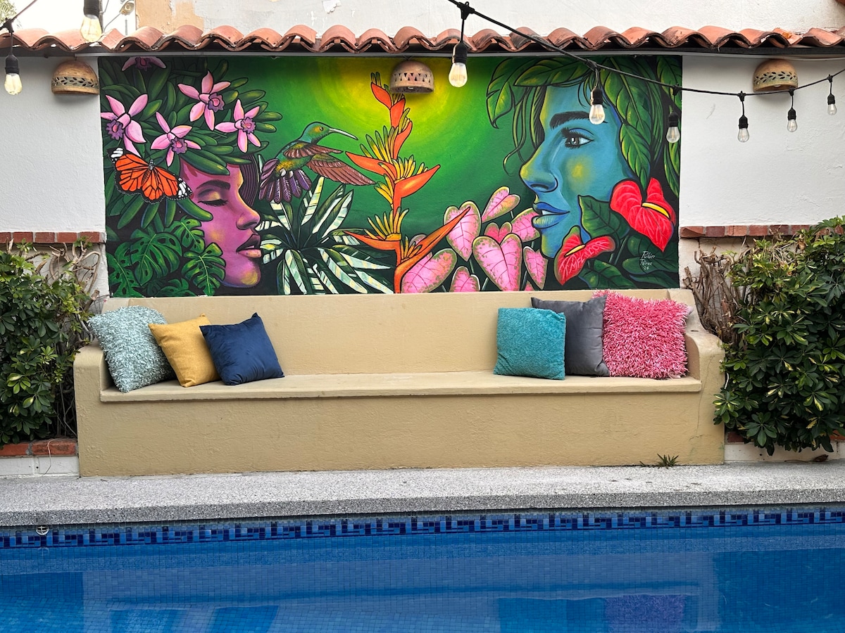 A vibrant mural depicting a woman's face and tropical foliage is displayed on a garden wall. A long, beige bench is positioned in front, adorned with colorful pillows. Decorative string lights hang overhead, illuminating the tranquil setting near a swimming pool.