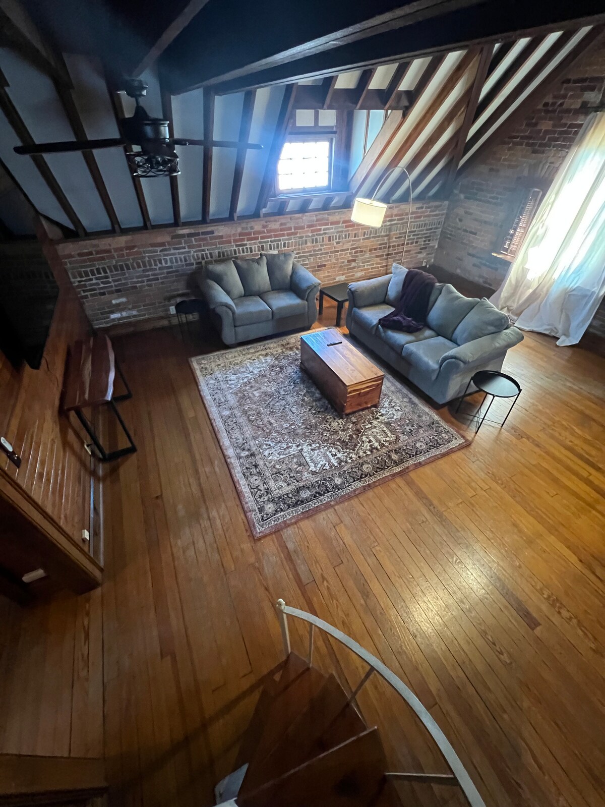 The living space is characterized by exposed brick walls and wooden beams. A pull-out queen size sofa bed and a loveseat are arranged around a central coffee table. A decorative area rug lies on the hardwood floor, with natural light streaming in through the windows.