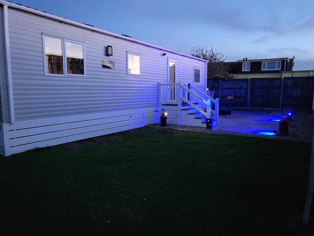 A mobile home exterior is shown, featuring white paneling and multiple windows. Blue lighting highlights the steps leading up to the entrance. A spacious outdoor area, with a patio and grass, is visible, creating an inviting environment for relaxation.