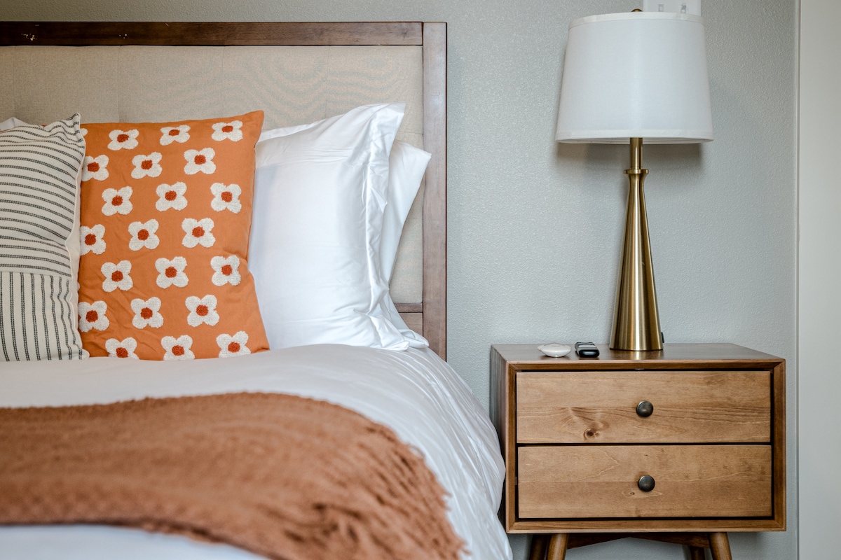 A cozy bedroom setting features a spacious bed adorned with white linens and decorative pillows, including an orange floral-patterned accent. A light wood nightstand with two drawers and a modern lamp offers a warm touch, against softly painted walls that create a serene atmosphere.