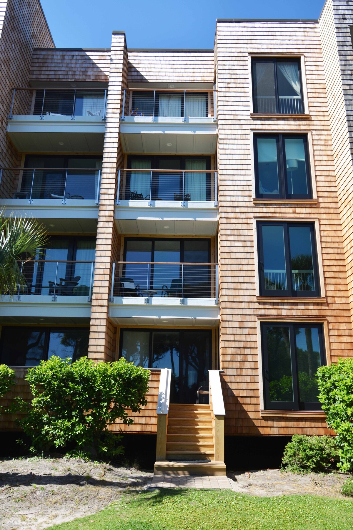 The exterior of a multi-story beach condo is displayed, showcasing wooden siding and large windows. A set of stairs leads to the entrance, framed by lush greenery. Balconies are visible on each level, offering outdoor space and views.