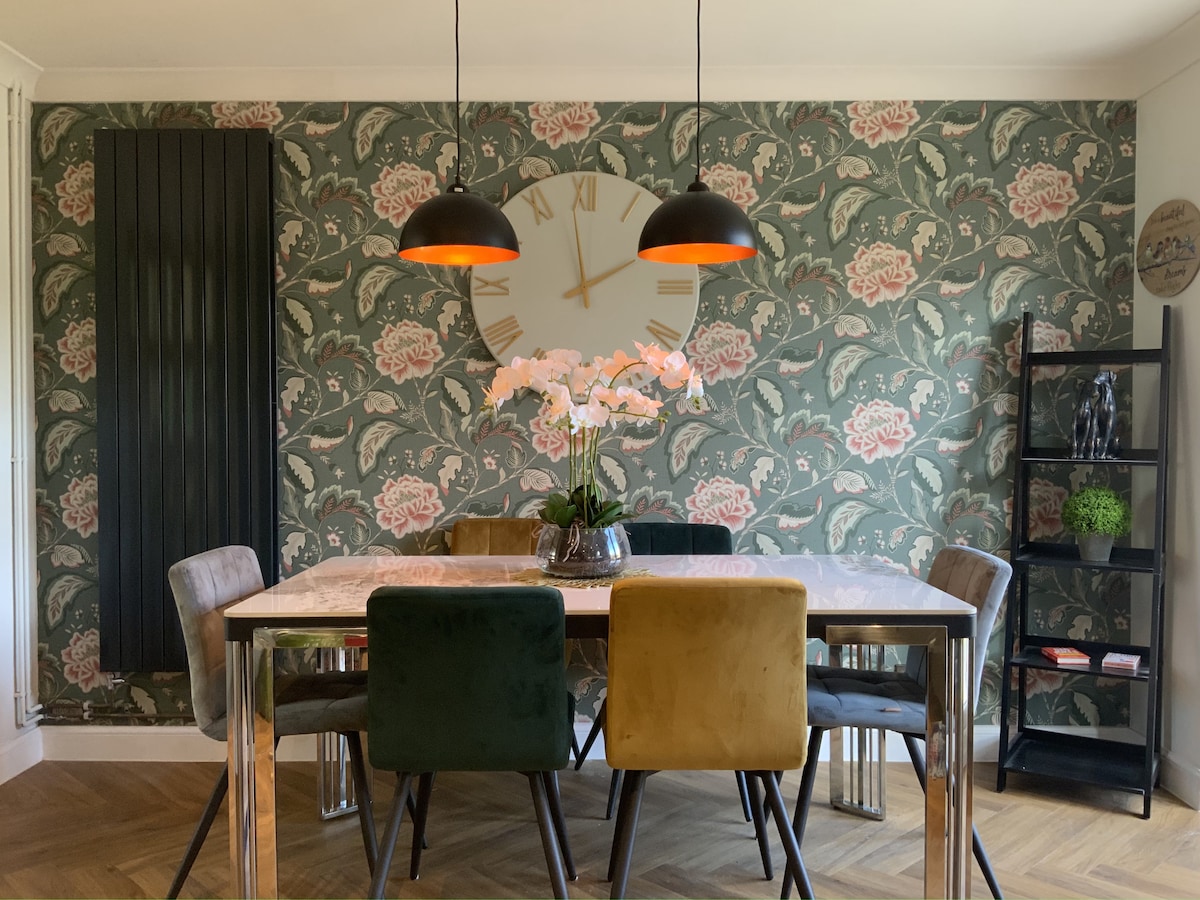 A dining area features a marble-topped table surrounded by upholstered chairs in various colors. A stylish clock adorns the floral-patterned wallpaper. An arrangement of white flowers in a vase adds a touch of elegance, complemented by warm pendant lighting overhead.