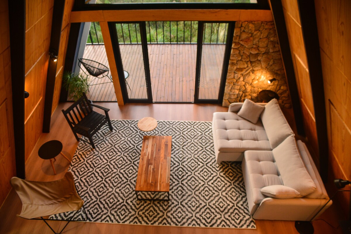 An open living area features a light gray sofa, a wooden coffee table, and a patterned area rug. Large glass doors provide views of the deck and surrounding nature. A comfortable chair and a small side table complete the inviting space, enhanced by warm lighting elements.