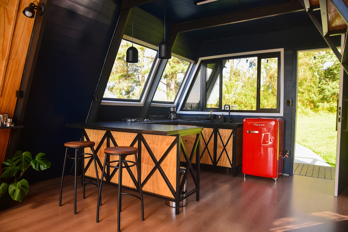 A modern kitchen is presented with large windows allowing natural light to fill the space. The island features a black countertop and wood-accented design, accompanied by high stools. A striking red refrigerator stands against a wall, adding a pop of color to the serene environment.