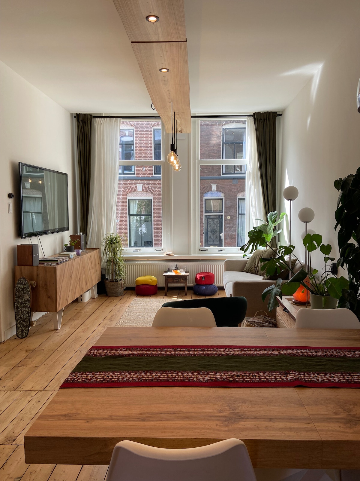 The living area features large windows allowing natural light to fill the space. A wooden dining table is accented by a colorful table runner. A comfortable seating arrangement is complemented by greenery and decorative elements, providing a relaxing ambiance.