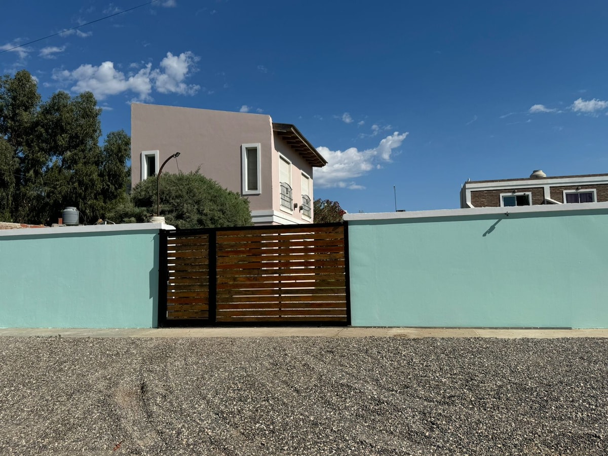 A wooden gate is set against a pastel turquoise wall, leading into a spacious outdoor area. A light-colored two-story building, visible behind the gate, features a balcony with decorative railings. The clear blue sky helps to create an open and inviting atmosphere.