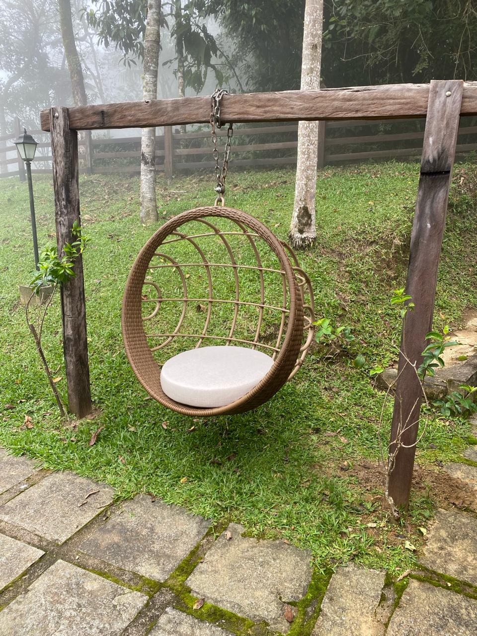 A suspended round wicker chair is positioned on a grassy area, surrounded by lush greenery and trees. The chair's soft cushion is visible, inviting relaxation. A stone pathway leads toward the chair, enhancing accessibility to this serene outdoor space.